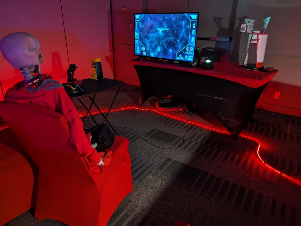 A plastic skeleton in a hoodie sitting in a gamer setup with desk, monitor, and peripherals.